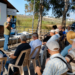 An outdoor agricultural presentation takes place beside a wooden shelter, where a speaker stands in front of a projected presentation and addresses an audience seated on plastic chairs. The event is held in a rural setting with trees, fields, and parked vehicles visible in the background.